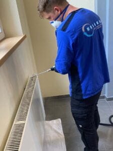 Eine Person in blauem Hemd, schwarzer Hose und Schutzmaske reinigt mit einem Werkzeug einen Heizkörper in einem Raum. Der Heizkörper ist an einer Wand mit beigem Gips montiert, auf dem Boden darunter ist ein weißes Tuch ausgebreitet.